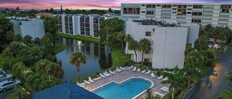 A hotel with a swimming pool, a parking area, and a view of the ocean at sunset.