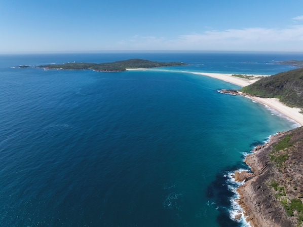 Experience the serene coastal landscape with gentle waves lapping onto the shore, surrounded by lush greenery and rugged cliffs.