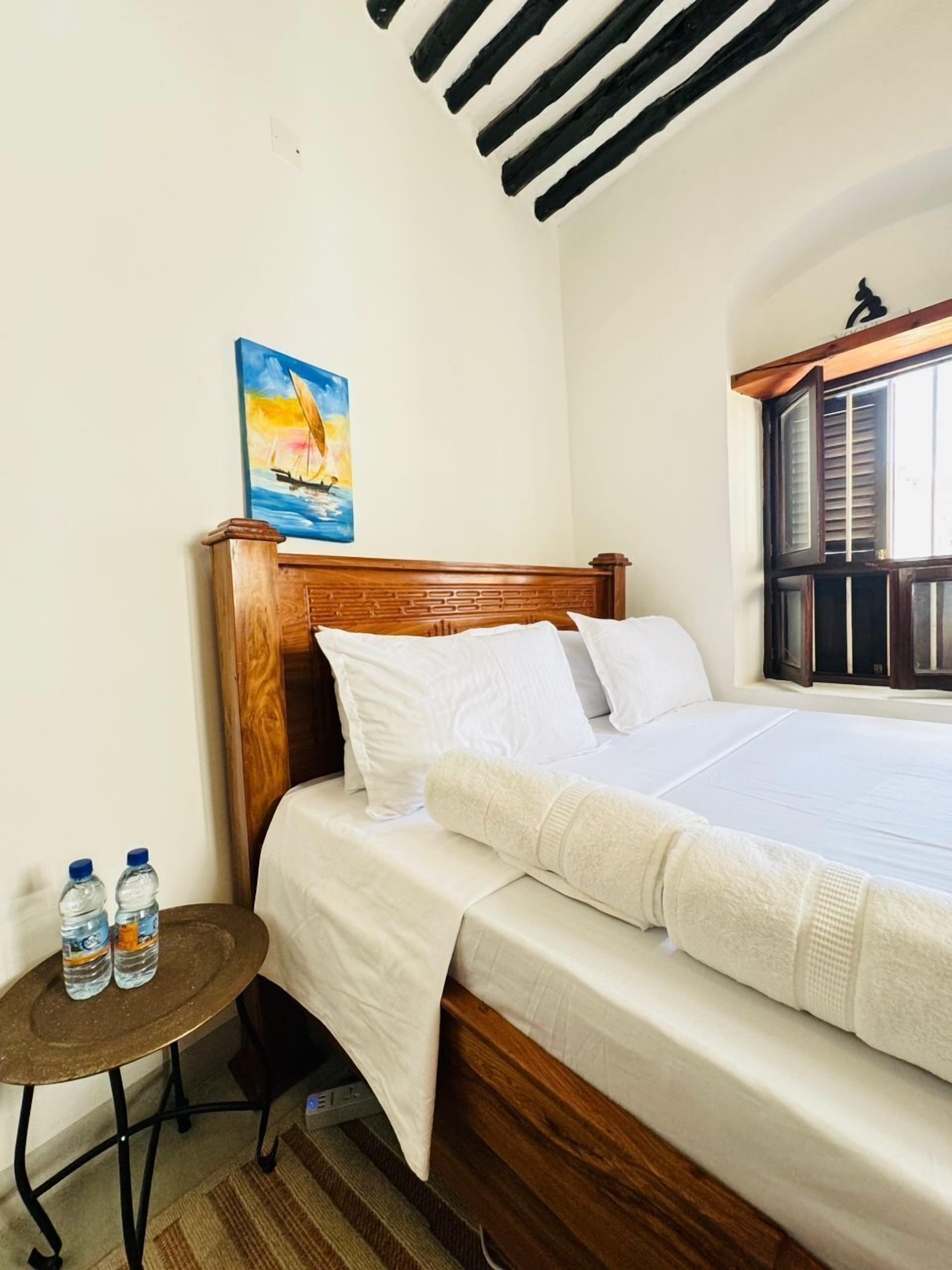 Una cama bien tendida con ropa blanca, un cabecero de madera y un cuadro de un velero en la pared. Hay una pequeña mesa redonda con dos botellas de agua al lado de la cama.