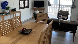 Zona pranzo con tavolo e sedie in legno, televisore su supporto e balcone con vista su vegetazione.