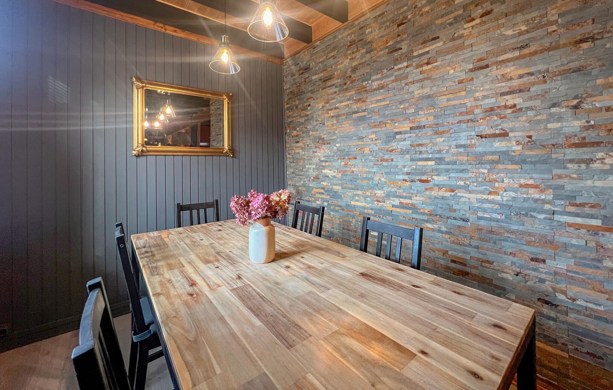 A dining area with a wooden table, chairs, a vase with flowers, a mirror, and a stone wall.