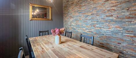 A dining area with a wooden table, chairs, a vase with flowers, a mirror, and a stone wall.