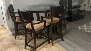A dining area with a round glass table and four chairs, a flat-screen TV, and a lamp.
