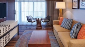 A hotel room with a sofa, a coffee table, a TV, a dining table with chairs, and a large window with curtains.