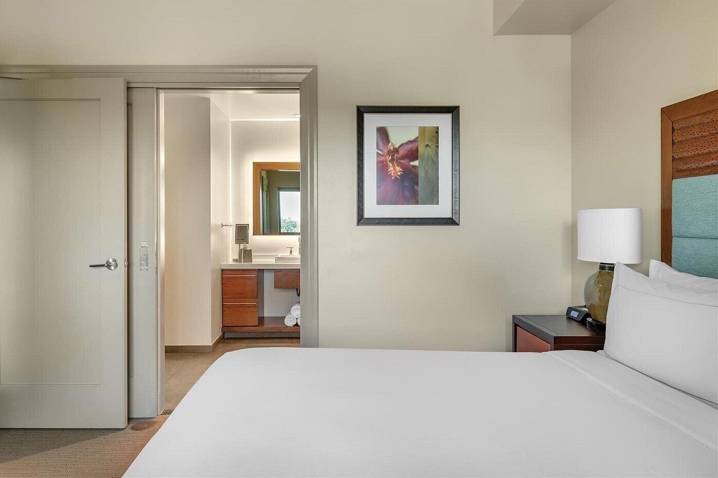 A hotel room with a bed, a nightstand with a lamp, a framed picture on the wall, and a bathroom visible through an open door.