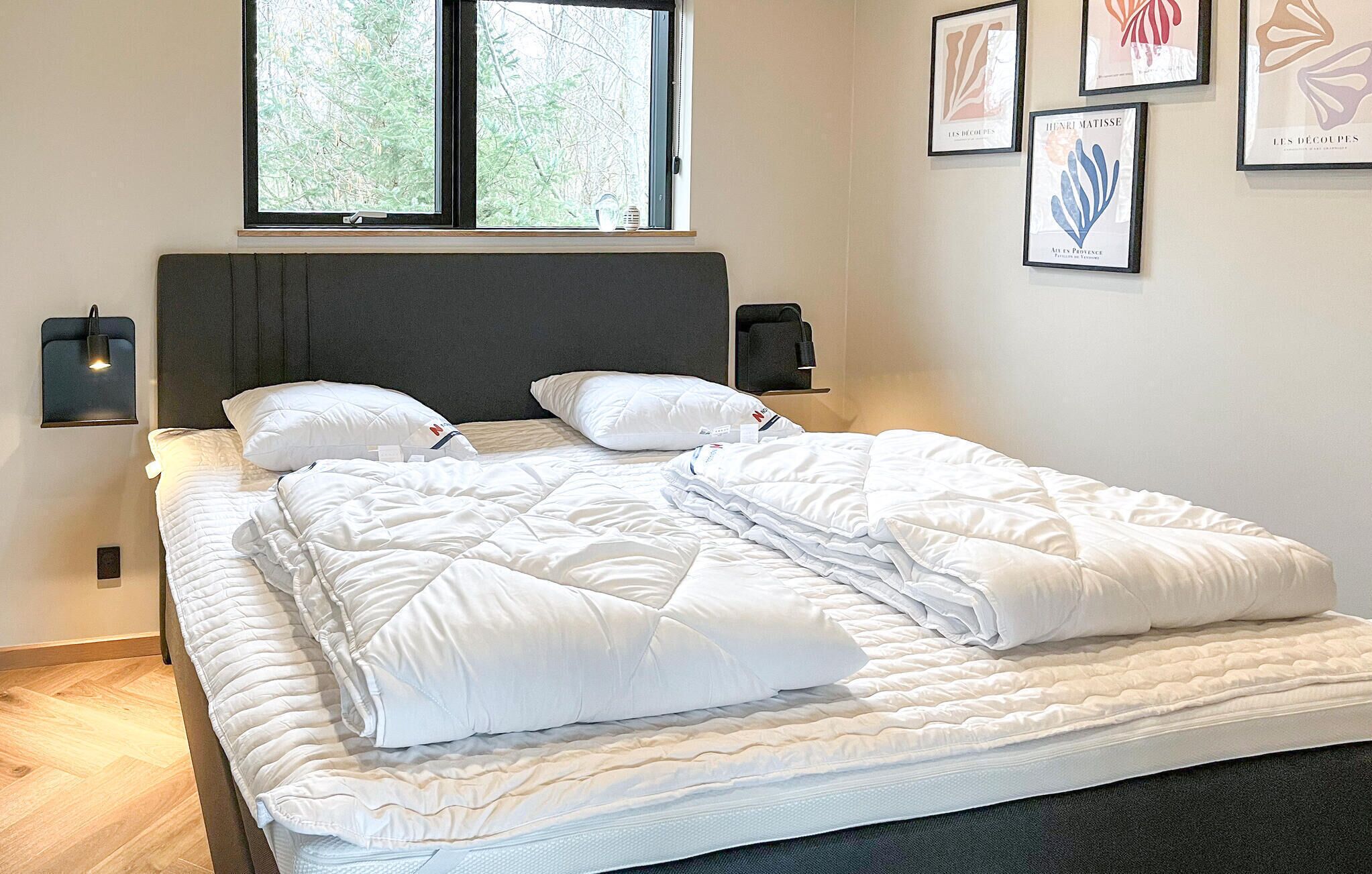 Cama bem arrumada com roupa de cama branca, cabeceira e janela com vista para árvores.