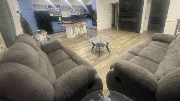 A modern living room with grey sofas, a glass coffee table, and a kitchen area with a bar counter and stools. - Charming 2-bedroom house with AC and WiFi in lovely Wanette (Wanette)