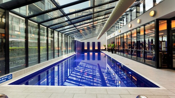 An indoor swimming pool with glass walls, a ceiling with a geometric design, and exercise equipment visible through the glass.