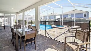Un patio techado con una mesa y sillas de vidrio, una piscina y vistas a casas al fondo.