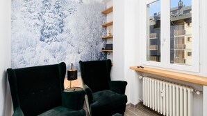 A room with a large window, two dark green armchairs, a radiator, and a wall-mounted shelf with a small plant.