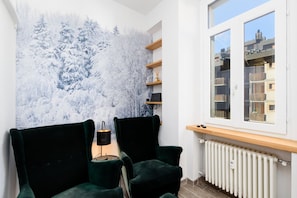 A room with a large window, two dark green armchairs, a radiator, and a wall-mounted shelf with a small plant.