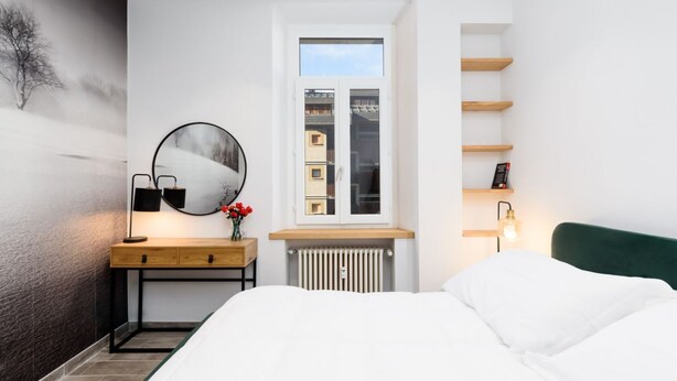 A bedroom with a bed, a bedside table with a lamp, a mirror, a window, and a radiator.