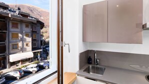 A modern kitchen with a sink and cabinets, a window overlooking parked cars and buildings, and a view of a hilly landscape.