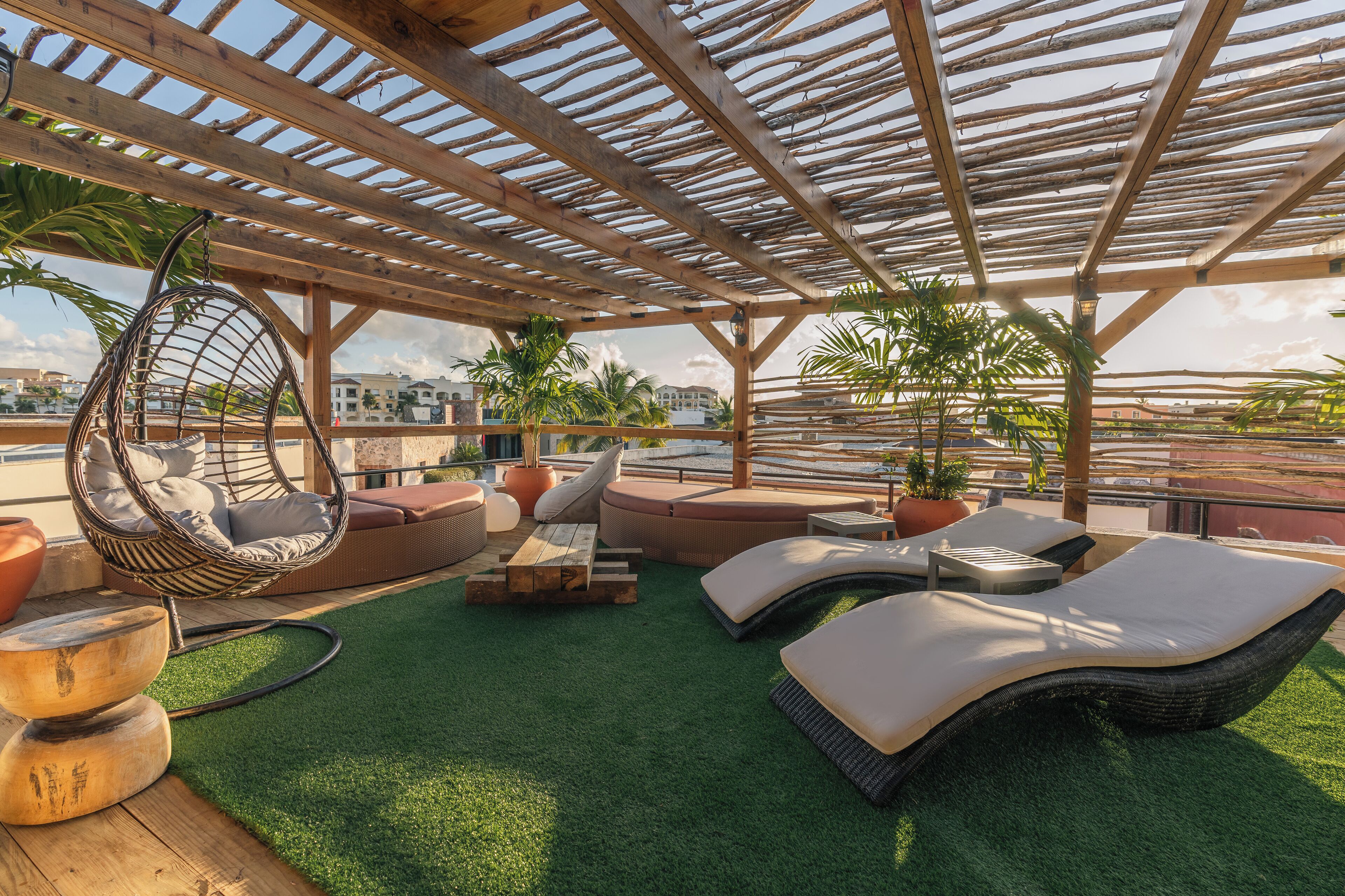 A rooftop lounge with a wooden pergola, hanging chairs, sofas, and artificial grass.