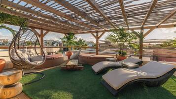 A rooftop lounge with a wooden pergola, hanging chairs, sofas, and artificial grass.