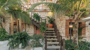 Una escalera de madera que conduce a un edificio de piedra y ladrillo con un jardín tropical y palmeras.