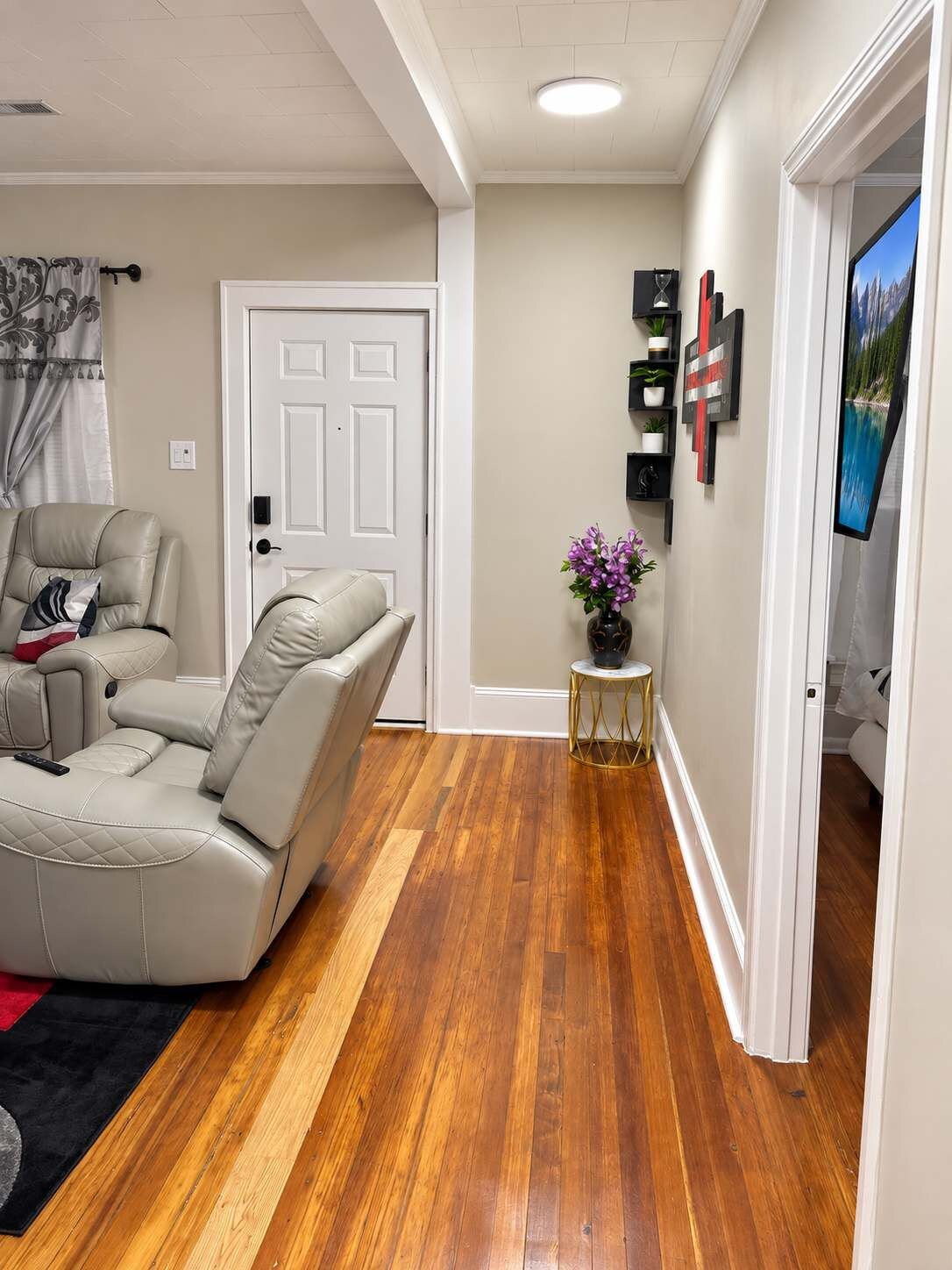 A living room with a beige sofa, a small round table with a vase of flowers, and a television on the wall.