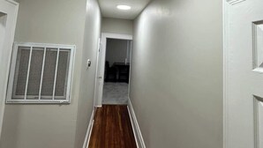 Un pasillo estrecho con paredes blancas y suelo de madera. A la derecha, una puerta blanca y, a la izquierda, una rejilla de ventilación. El pasillo da a una habitación con suelo oscuro.