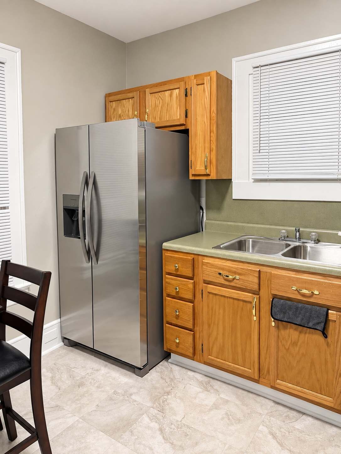 A kitchen with a stainless steel refrigerator, wooden cabinets, a countertop, and a window with blinds.