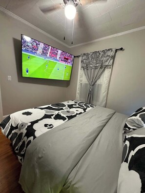 Ein Schlafzimmer mit einem Bett, einem Deckenventilator, einem an der Wand befestigten Fernseher und einem Fenster mit Vorhängen.