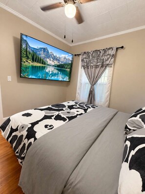 Un dormitorio con cama, ventilador de techo, televisor montado y ventana con cortinas.