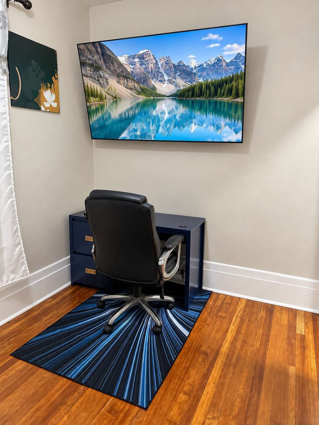 Een kamer met een grote televisie aan de muur, waarop een bergmeer te zien is, een zwarte bureaustoel, een bureau met lades en een blauw tapijt met patroon op een houten vloer.
