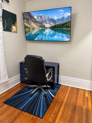 Habitación con una televisión grande montada en la pared que muestra una escena de un lago en la montaña, una silla de escritorio negra, un escritorio con cajones y una alfombra azul estampada sobre un piso de madera.