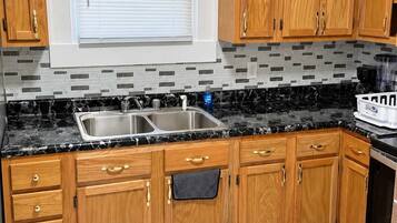 A kitchen with wooden cabinets, a black marble countertop, a double sink, and a window with blinds.