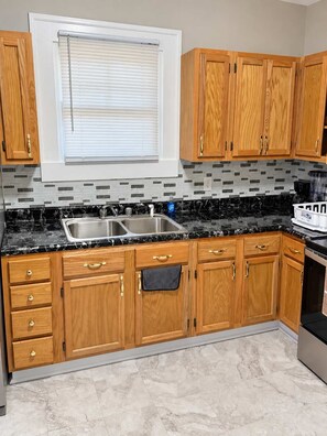 A kitchen with wooden cabinets, a black marble countertop, a double sink, and a window with blinds.