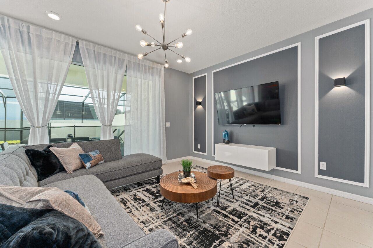 A modern living room with a grey sofa, a round coffee table, a TV mounted on the wall, and a large window with sheer curtains.