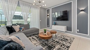 Ein modernes Wohnzimmer mit einem grauen Sofa, einem runden Couchtisch, einem an der Wand montierten Fernseher und einem großen Fenster mit transparenten Vorhängen.
