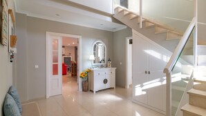A spacious hallway with a staircase leading to an upper floor. A white cabinet with a mirror and a vase of sunflowers is visible.