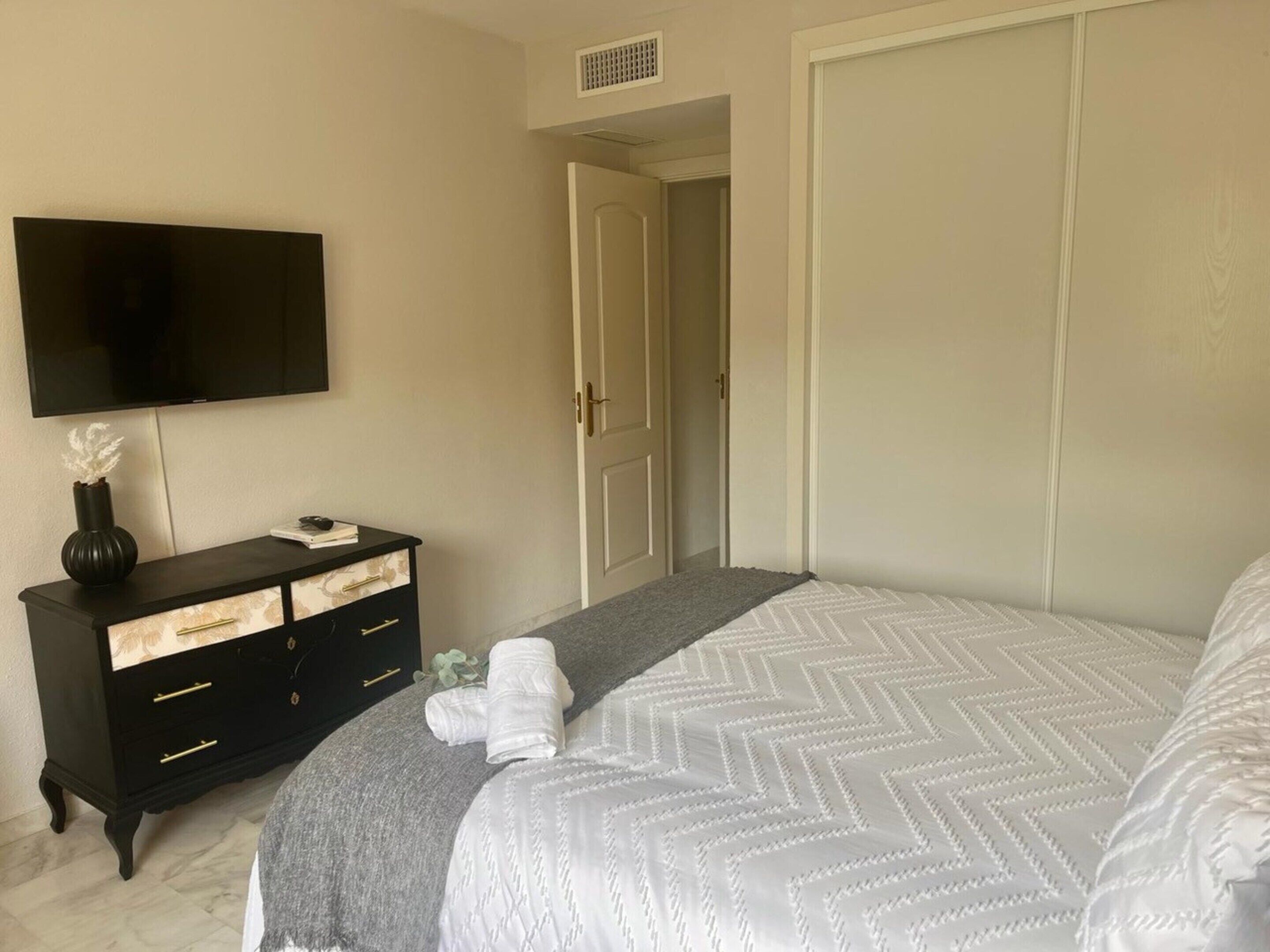 A bedroom with a bed, a TV mounted on the wall, a dresser, and a door leading to another room.