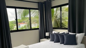 Une chambre moderne avec un grand lit, une table de chevet, une lampe et une vue sur les palmiers par les fenêtres.