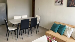 Un coin repas avec une table et des chaises blanches, un canapé beige agrémenté de coussins turquoise et une table basse blanche ornée de fleurs.