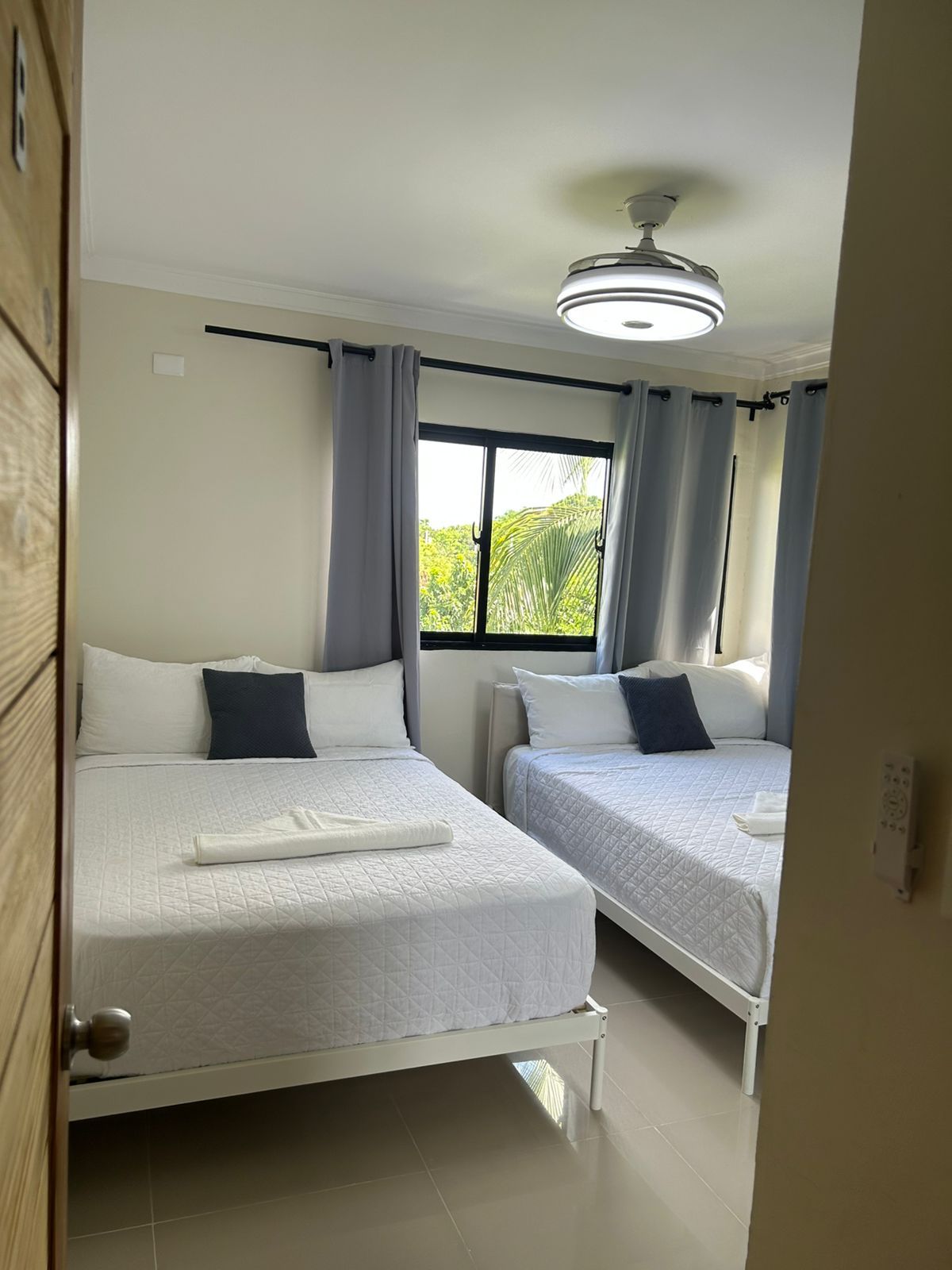 Habitación de hotel con dos camas, una ventana con cortinas, ventilador de techo y vista a zonas verdes.