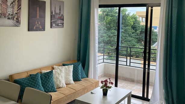 Un salon moderne avec un canapé, une table basse et une table à manger. Trois tableaux encadrés ornent le mur, un luminaire est suspendu au plafond, et la fenêtre offre une vue sur la verdure.