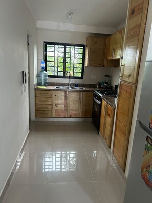 A kitchen with wooden cabinets, a stainless steel sink, a stove, and a window with a view of greenery. - FREE CLEANING / NO FEES / Comfortable Apartment Near AILA Airport & Boca Chica (Santo Domingo Este)