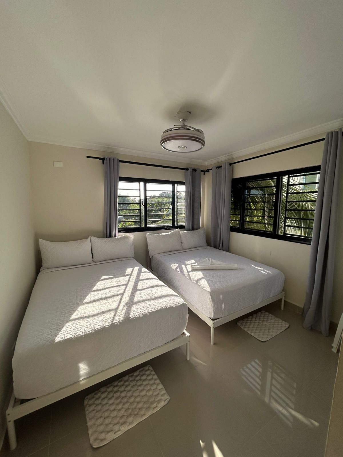 Ein Schlafzimmer mit zwei Betten, einem Deckenventilator und großen Fenstern mit Vorhängen.