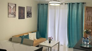 Un salon moderne avec un canapé, une table à manger et des chaises. On y trouve trois tableaux accrochés au mur, un ventilateur de plafond et une fenêtre avec des rideaux.