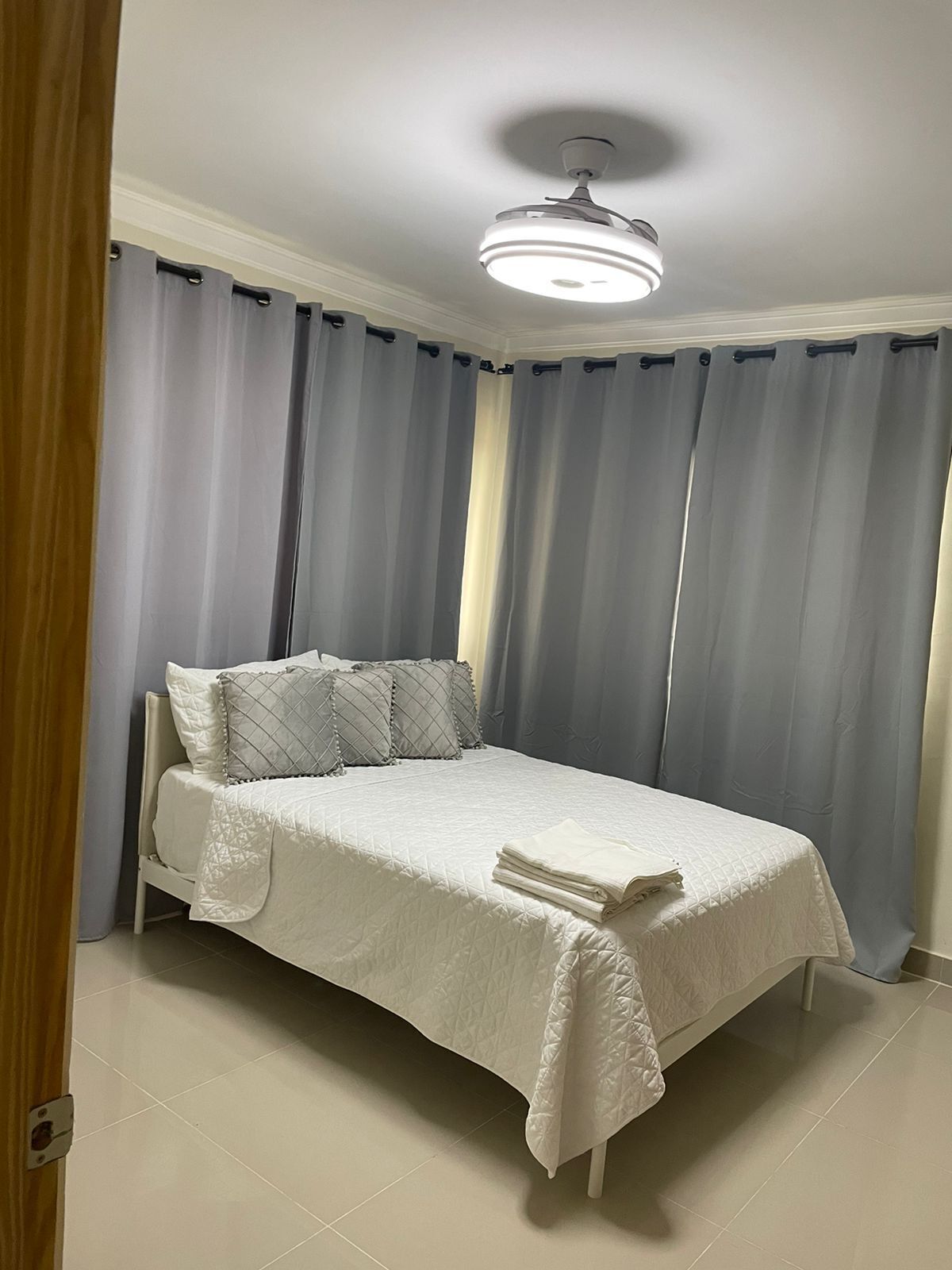 A neatly made bed with a white bedspread and pillows, a ceiling fan, and light blue curtains.