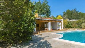 Zona de piscina con área de estar techada, una piscina y vegetación circundante.