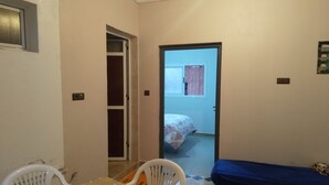 Una habitación pequeña y funcional con una mesa de comedor y sillas, una cama individual y una puerta que da a otra habitación.