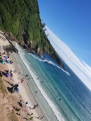 Een strand met mensen die zwemmen en wandelen langs de kust, omgeven door weelderige begroeiing en een helderblauwe lucht.