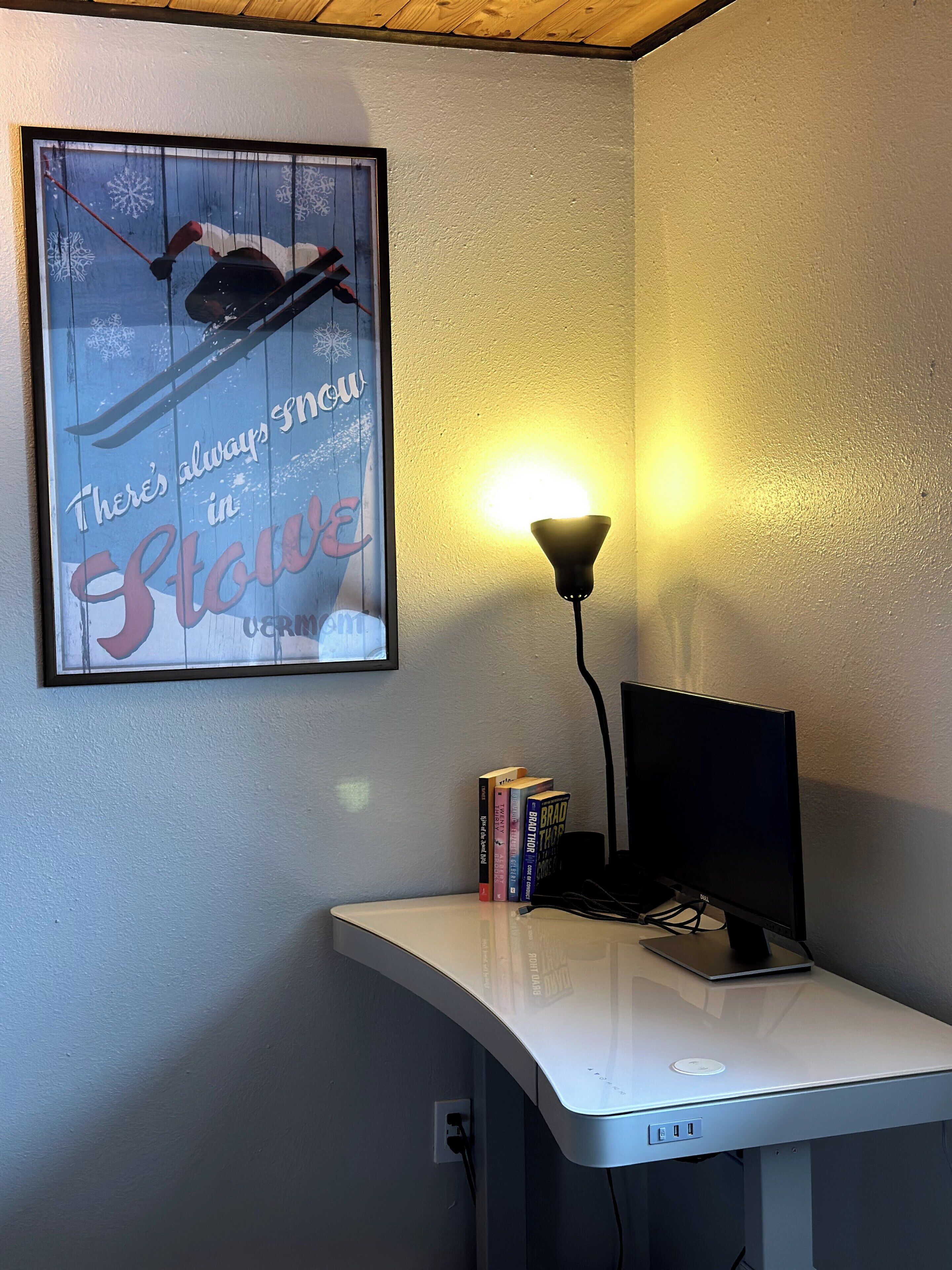 A desk with a laptop, a lamp, and books. A framed poster with a winter theme is on the wall.