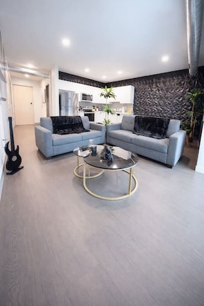 A modern living room with a glass coffee table, grey sofas, and a kitchen in the background. - The Green & Onyx Escape (Des Moines)