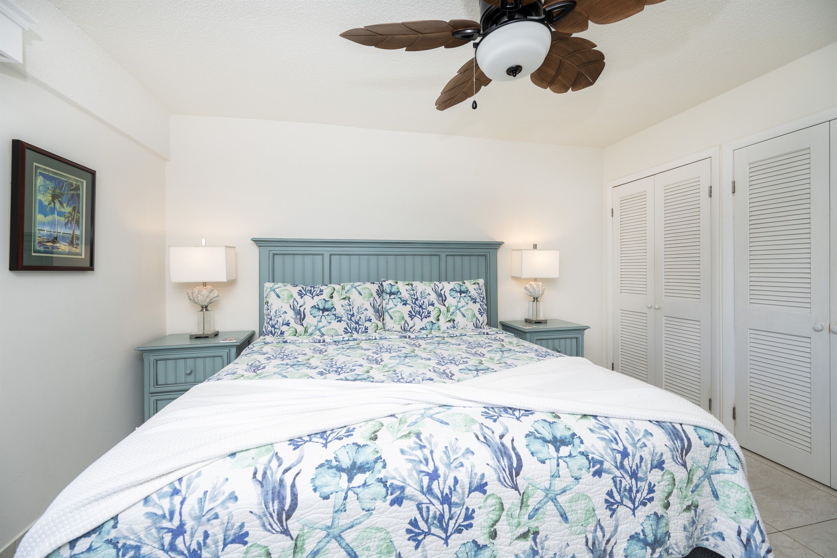 A bedroom with a bed, two bedside tables with lamps, a ceiling fan, and white closets.