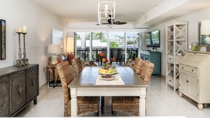 A dining area with a wooden table, wicker chairs, and a fruit bowl. There is a ceiling fan, a cabinet with decorative items, and a television on a stand. - Beachfront, Ocean Views, 2 bed, Poolside, Gym (St. Thomas)