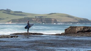 A person standing on rocks by the sea with a surfboard, overlooking a coastline with cliffs.