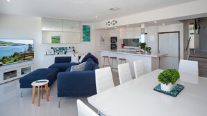 A modern living room with a large TV, blue sofas, a dining table with white chairs, and a kitchen area with a fridge and bar stools.